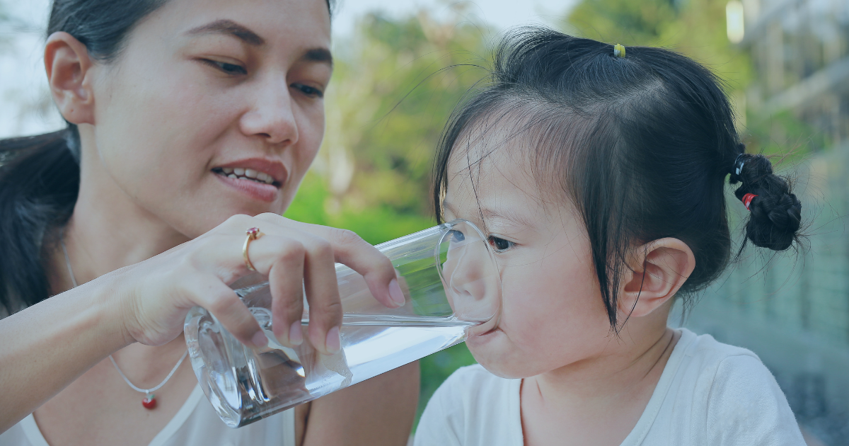 A woman helps a young child drink water from a glass outdoors.