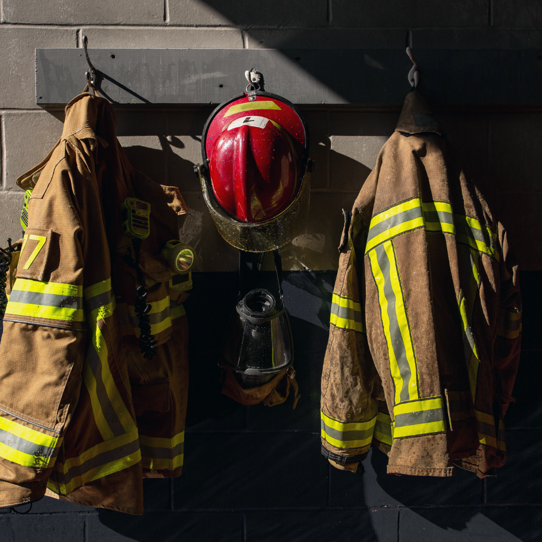 Firefighter gear hangs on a brick wall, including a red helmet and two jackets with reflective stripes.