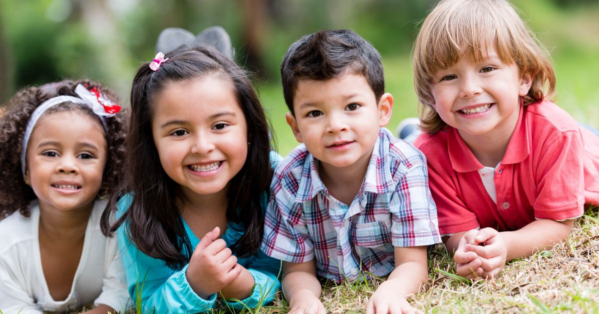 Four smiling children lie on grass, facing the camera.