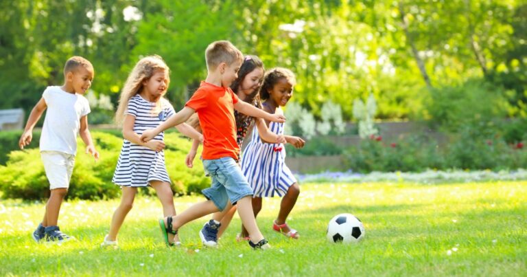 Five children play soccer together on a grassy field in a park on a sunny day, with trees in the background.