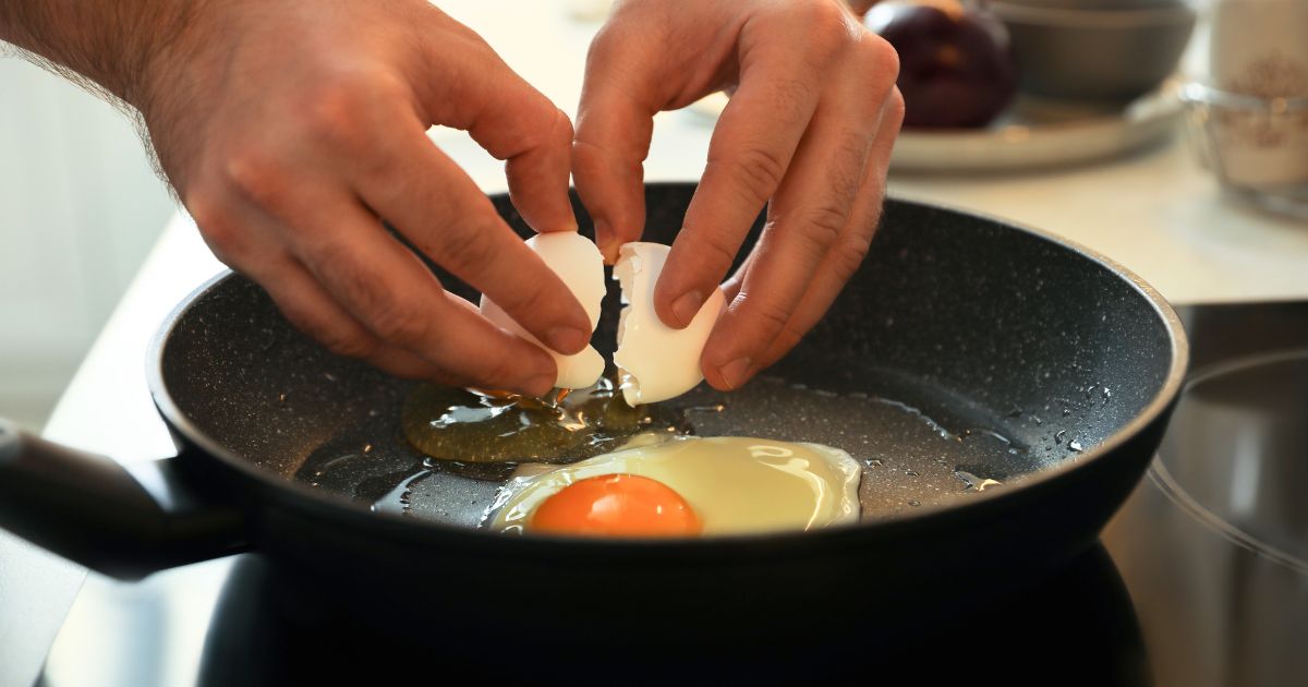 Hands crack an egg into a sizzling black frying pan, with the yolk and whites spreading.
