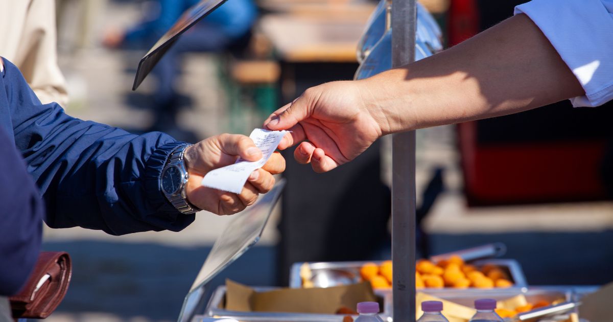 A hand in a white sleeve exchanges a receipt with another hand in a blue jacket at an outdoor food stall, with trays of food visible in the background.