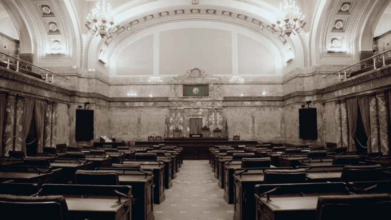 State legislative chamber with ornate ceiling, chandeliers, and marble walls. Rows of desks face a central podium.