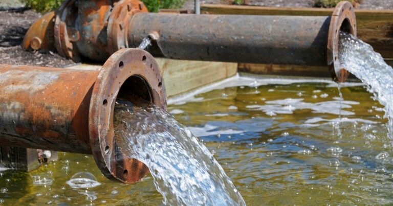Two large, rusty pipes discharge clear water into a pond, creating ripples.